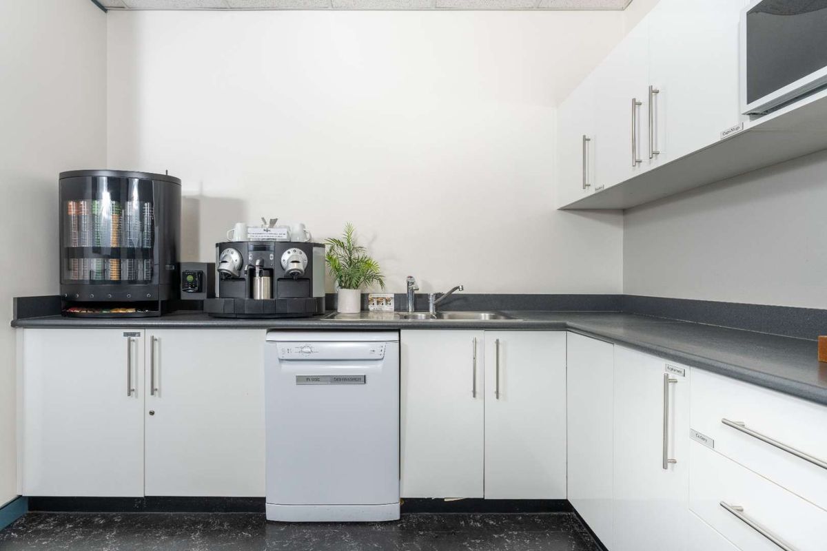 A functional office kitchenette featuring white cabinetry, dark laminate countertops, a stainless steel sink, and integrated appliances including a dishwasher and microwave.