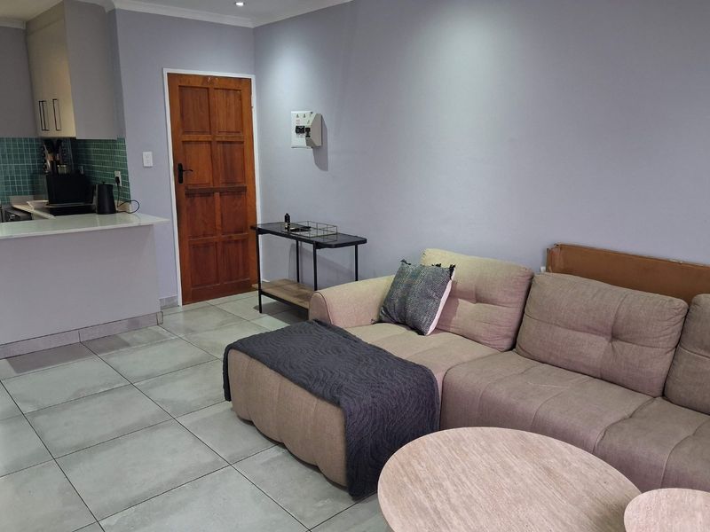 Open-plan living area featuring tiled flooring, a compact kitchen with tiled backsplash, and a solid timber door leading to an adjacent space.