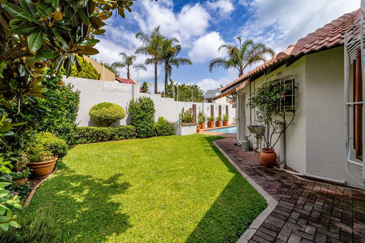 A well-maintained rear garden featuring a manicured lawn, brick-paved pathway, perimeter wall with security fencing, and a glimpse of the swimming pool area.