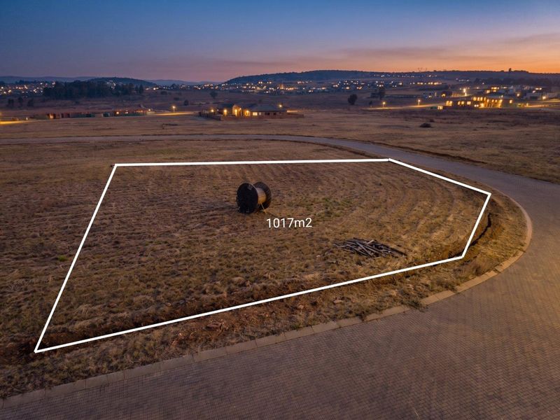 An aerial view of a 1017m2 vacant residential plot situated in a developed estate with paved road access and surrounding landscape views.