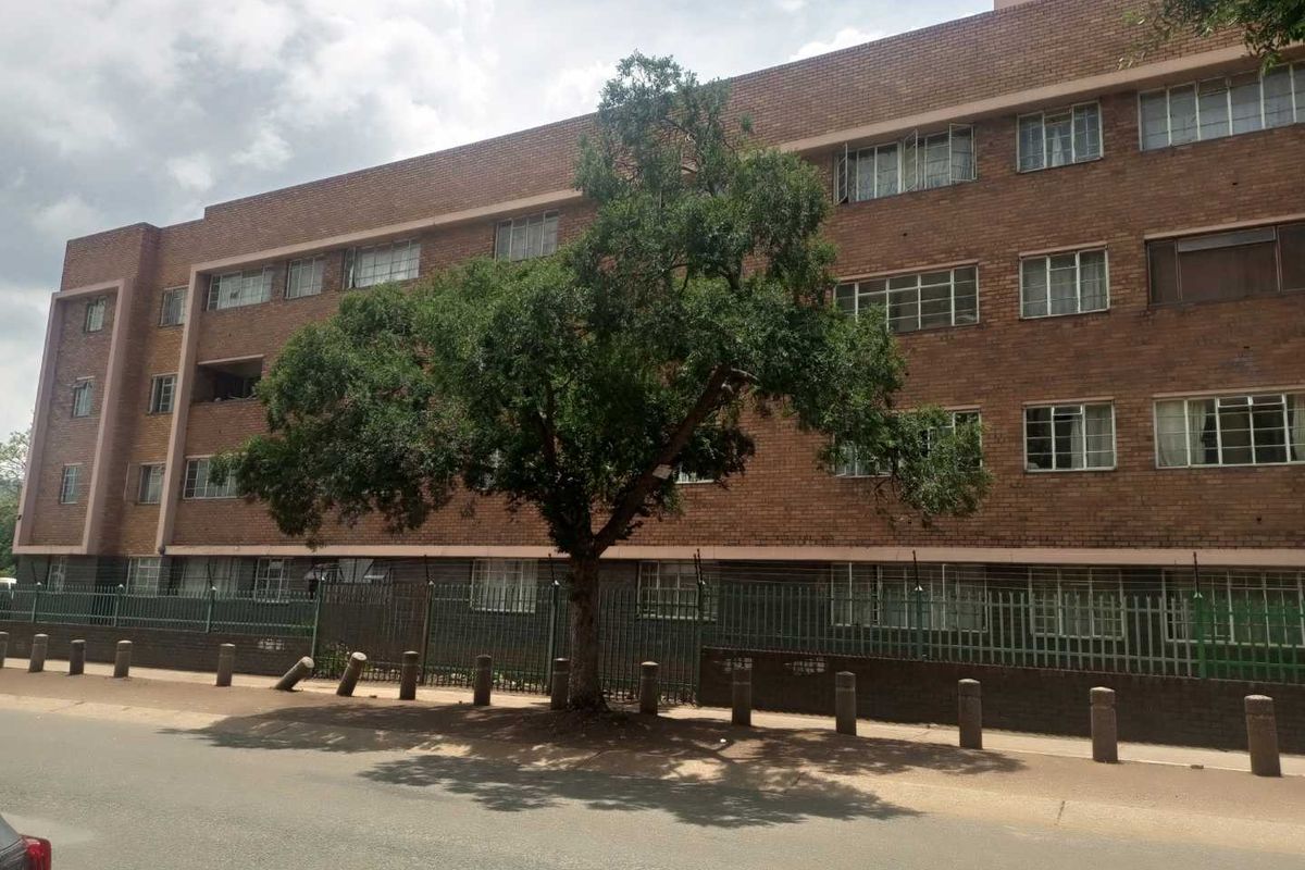 Street-level view of a multi-story brick apartment building featuring steel-framed windows and perimeter security fencing.
