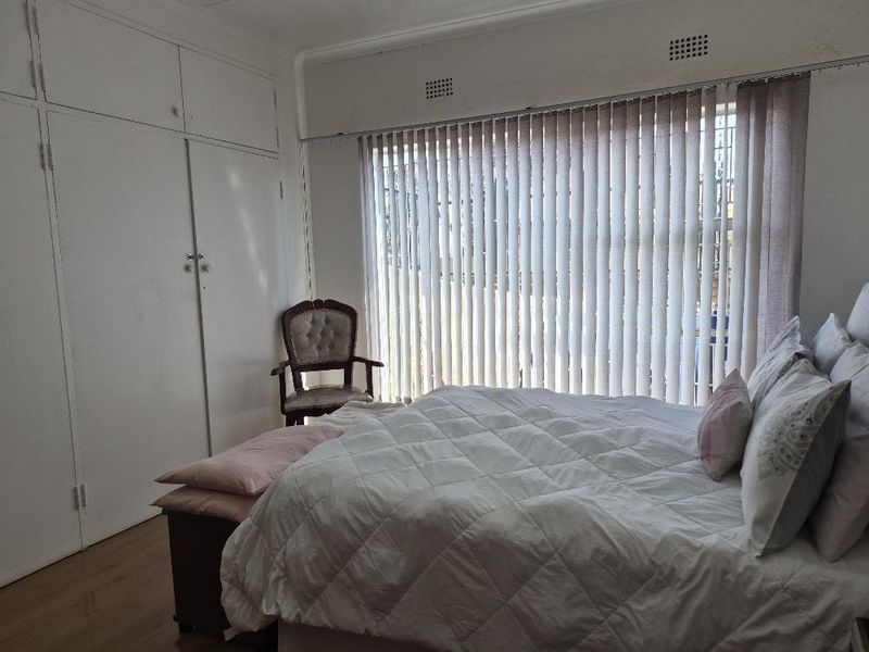 A bedroom featuring built-in cupboards, laminate flooring, and large windows fitted with vertical blinds, offering a functional and neutral living space.