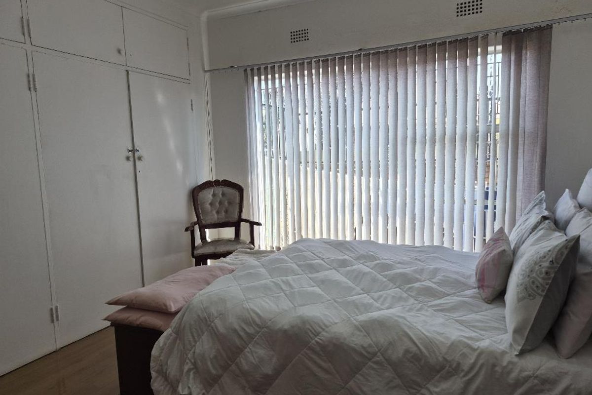 A bedroom featuring built-in cupboards, laminate flooring, and large windows fitted with vertical blinds, offering a functional and neutral living space.