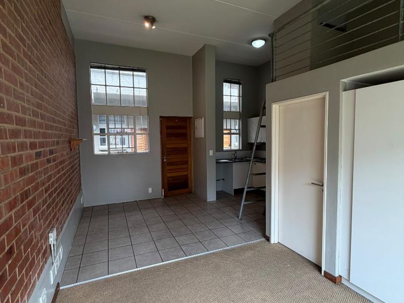 Open-plan living area featuring an exposed brick wall, mezzanine level with metal railing, kitchenette, and a mix of tiled and carpeted flooring.