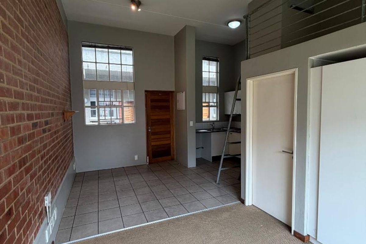 Open-plan living area featuring an exposed brick wall, mezzanine level with metal railing, kitchenette, and a mix of tiled and carpeted flooring.