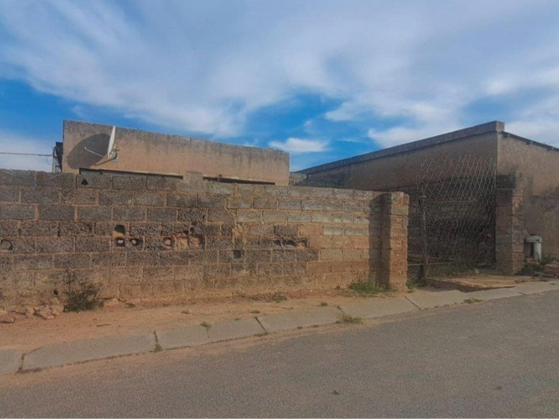 Street view of a property featuring a weathered masonry boundary wall and a metal gate, showing significant signs of wear and deterioration.
