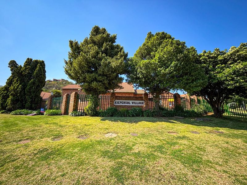 Front view of Kiepersol Village townhouse complex featuring a well-maintained lawn, mature trees, and brick boundary wall with security fencing.