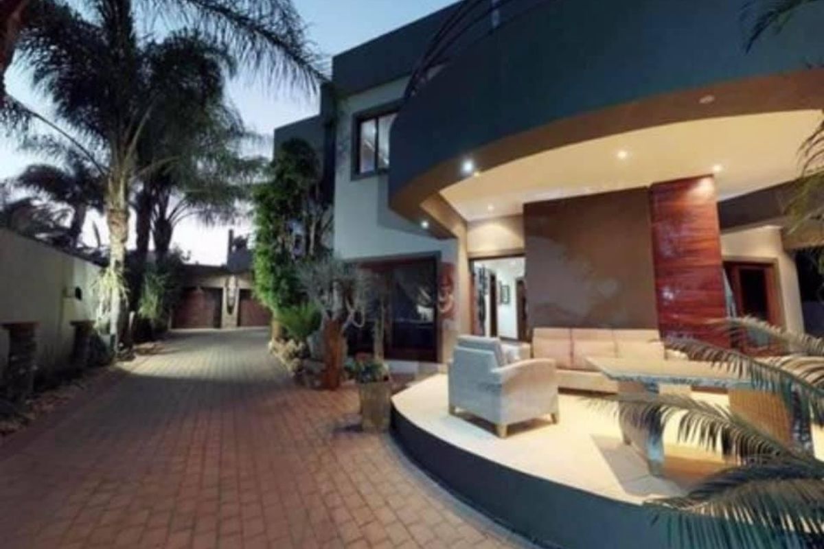 A modern covered patio area featuring a curved design, recessed lighting, and stone accents, overlooking a paved driveway and landscaped garden.