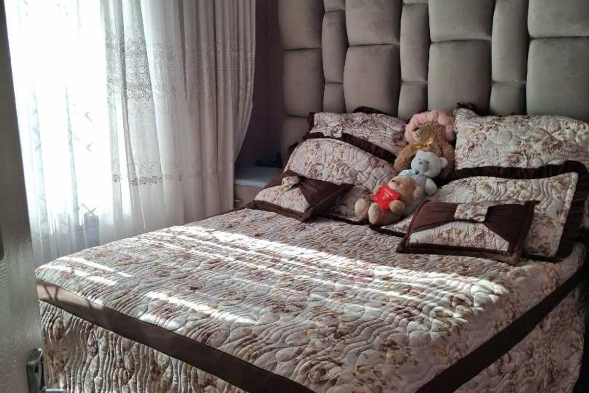 A cozy bedroom featuring a large upholstered headboard, ceiling cornice detailing, and tiled flooring, illuminated by natural light through draped windows.