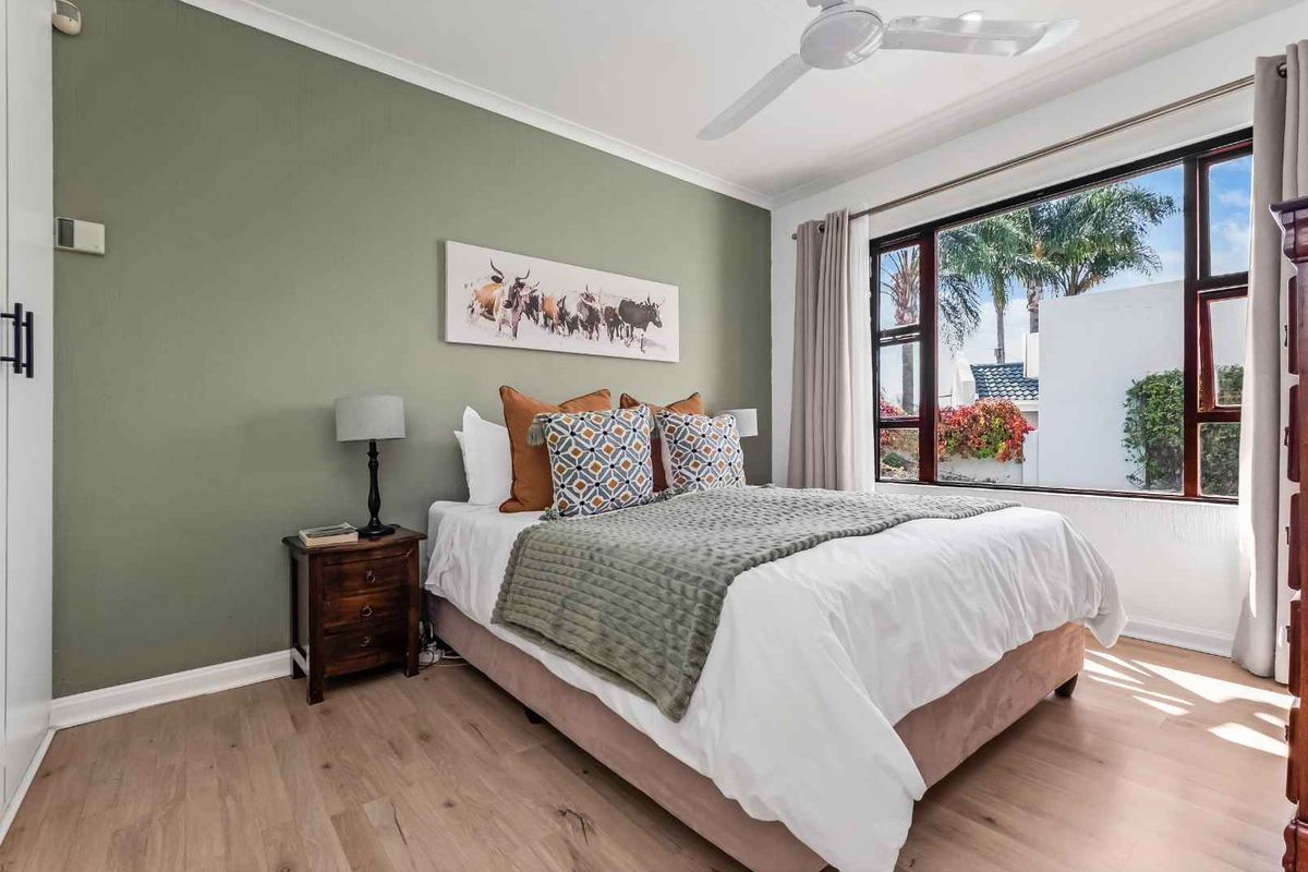 A well-lit bedroom featuring light wood-look flooring, a sage green accent wall, a ceiling fan, and a large window overlooking the garden.