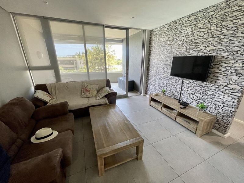 A bright living room featuring large sliding glass doors leading to a balcony, tiled flooring, and a decorative stone-effect feature wall.
