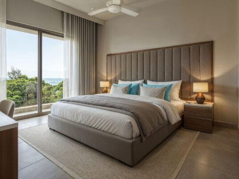 A modern, well-appointed bedroom featuring a large upholstered headboard, ceiling fan, and floor-to-ceiling sliding glass doors offering scenic coastal views.
