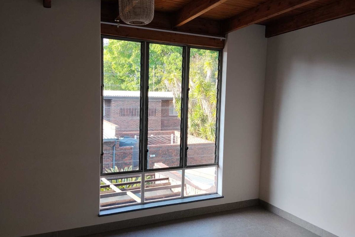 A bright, empty bedroom featuring exposed timber ceiling beams, large aluminium windows, and neutral tiled flooring, offering a clean and versatile living space.