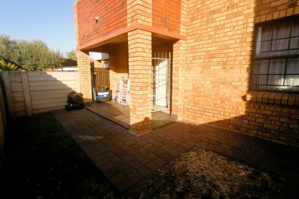 Rear exterior view of the apartment featuring a brick facade, a covered patio area, and a paved walkway leading to a small garden space.