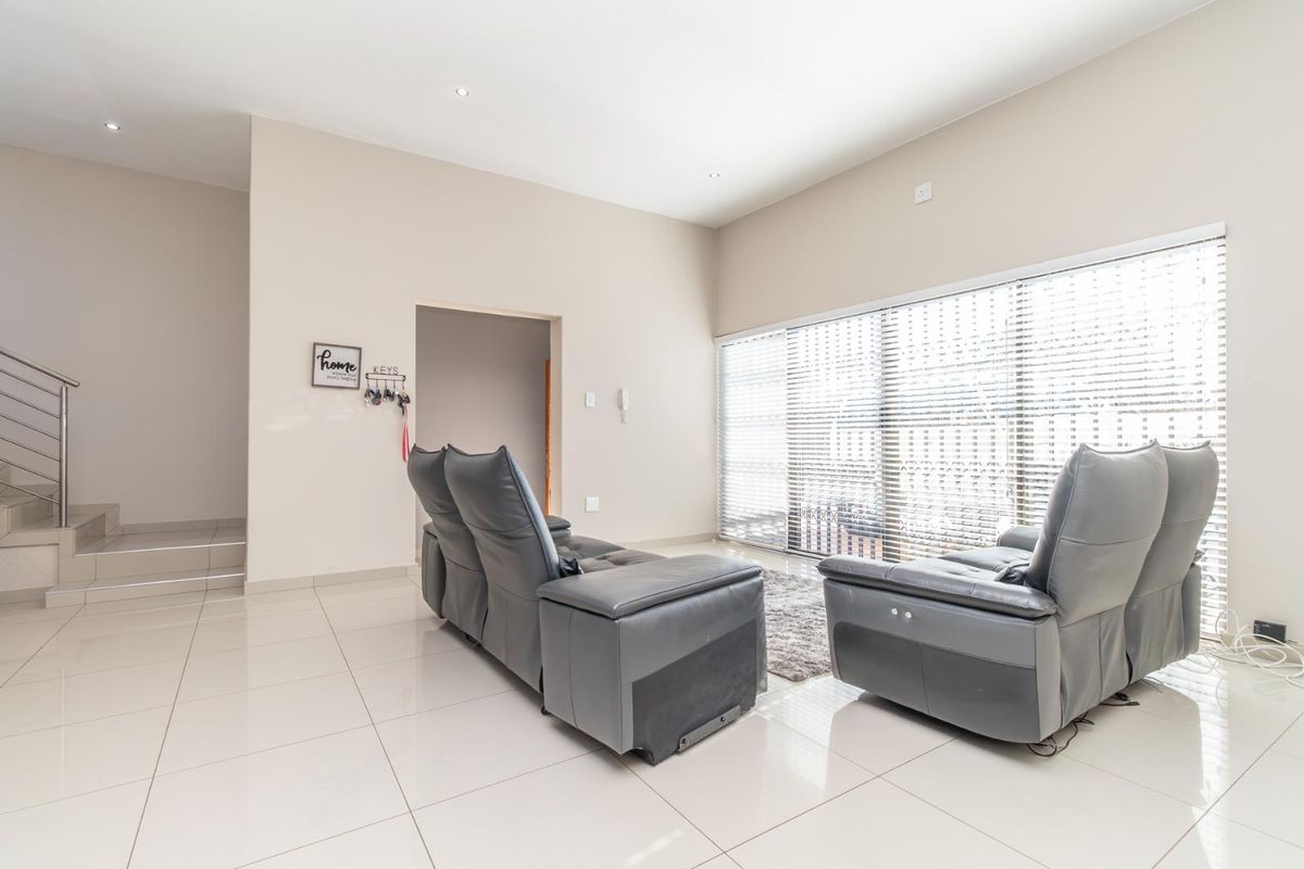 A bright and spacious living room featuring large windows with venetian blinds, polished tiled flooring, and a modern staircase with a stainless steel balustrade.