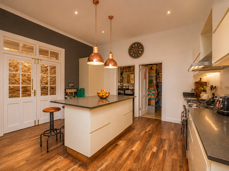 Modern kitchen featuring a central island with stone countertops, hardwood flooring, integrated appliances, and stylish pendant lighting, with access to a scullery area.