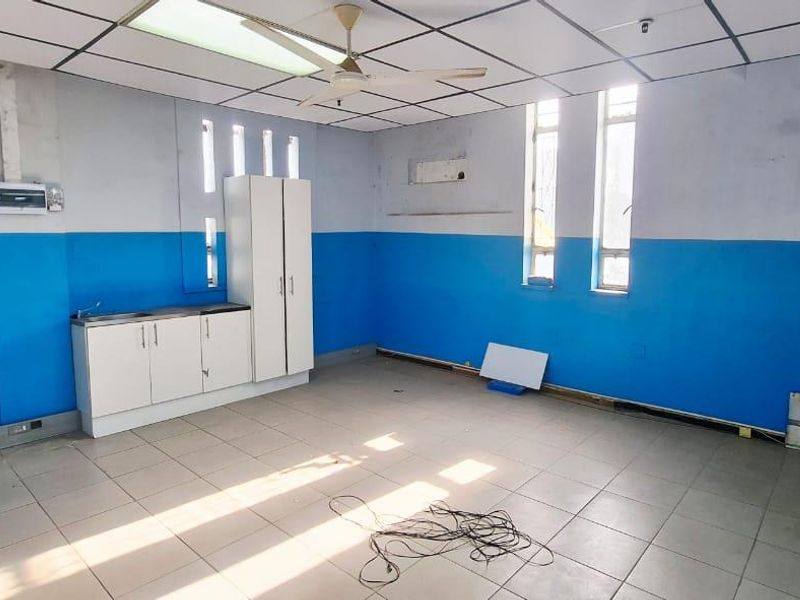 An empty office space featuring tiled flooring, a suspended ceiling with a fan, and a small built-in kitchenette unit with a sink.