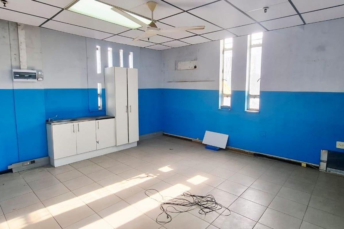 An empty office space featuring tiled flooring, a suspended ceiling with a fan, and a small built-in kitchenette unit with a sink.