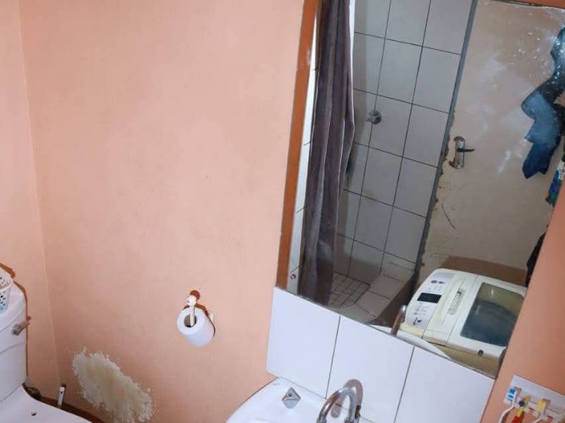 A compact bathroom featuring a pedestal basin, toilet, and shower area, currently showing signs of wall surface damage and wear.