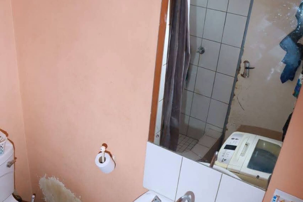 A compact bathroom featuring a pedestal basin, toilet, and shower area, currently showing signs of wall surface damage and wear.