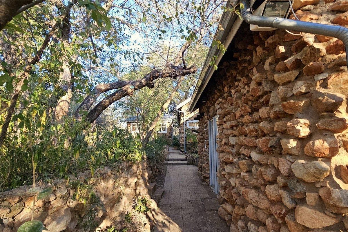 A rustic stone-clad exterior pathway leading alongside the house, surrounded by mature trees and established garden landscaping.