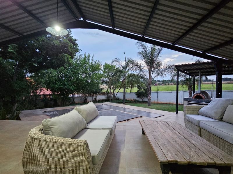 Spacious covered patio featuring tiled flooring and a built-in pizza oven, overlooking a swimming pool and landscaped garden area.