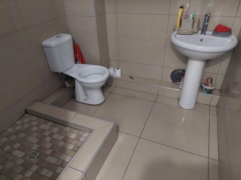 A functional bathroom featuring a pedestal basin, toilet, and a tiled shower base with neutral ceramic wall and floor tiling.