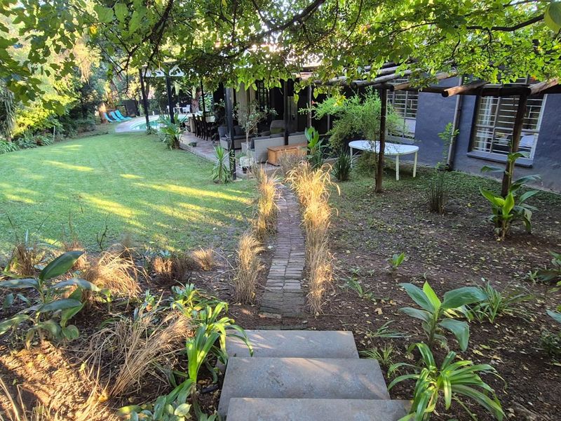 A spacious rear garden featuring a brick pathway, mature trees, a covered patio area, and a swimming pool in the background.