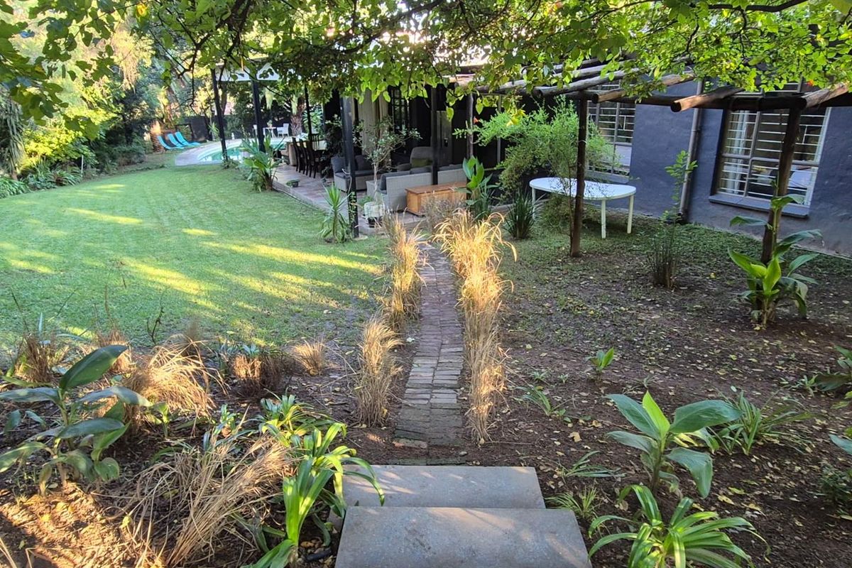 A spacious rear garden featuring a brick pathway, mature trees, a covered patio area, and a swimming pool in the background.