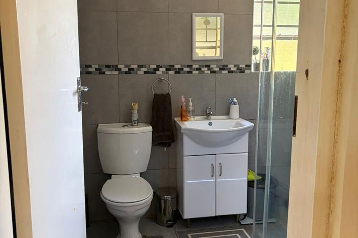 A compact bathroom featuring grey wall tiles, a white vanity unit with basin, a toilet, and a glass-enclosed shower.
