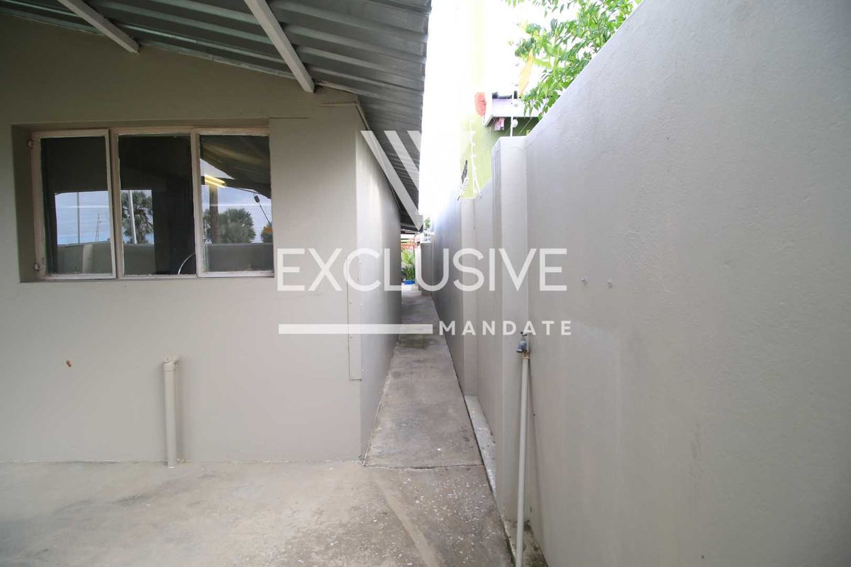A narrow side passage between the main building structure and the boundary wall, featuring a concrete pathway and an external water tap.