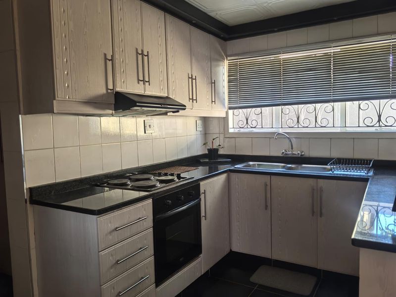 Functional kitchen featuring light-toned cabinetry, dark countertops, integrated electric hob and oven, tiled backsplash, and a double bowl sink with window security bars.