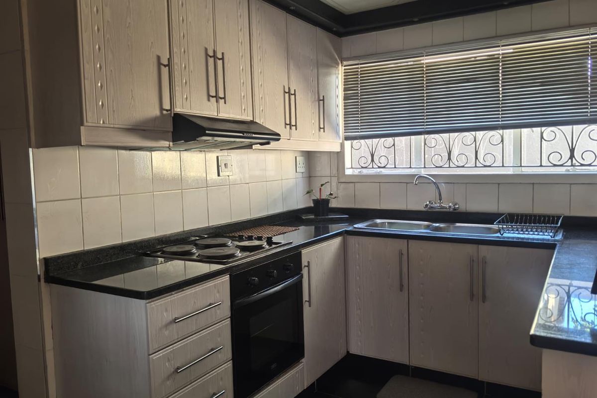 Functional kitchen featuring light-toned cabinetry, dark countertops, integrated electric hob and oven, tiled backsplash, and a double bowl sink with window security bars.