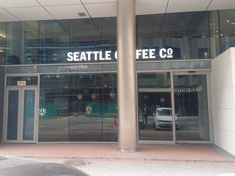 Modern office building exterior facade featuring a glass storefront entrance with metal structural columns and paved pedestrian access.