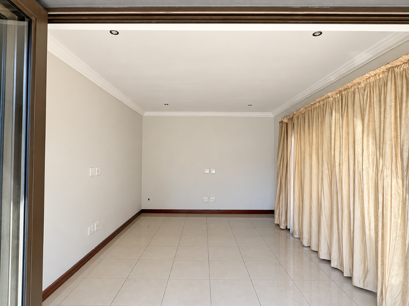A bright, empty living room featuring neutral tiled flooring, recessed downlighting, and large sliding glass doors with floor-to-ceiling curtains.