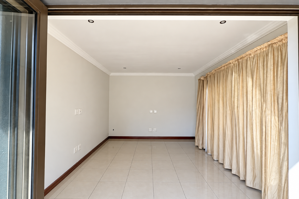 A bright, empty living room featuring neutral tiled flooring, recessed downlighting, and large sliding glass doors with floor-to-ceiling curtains.