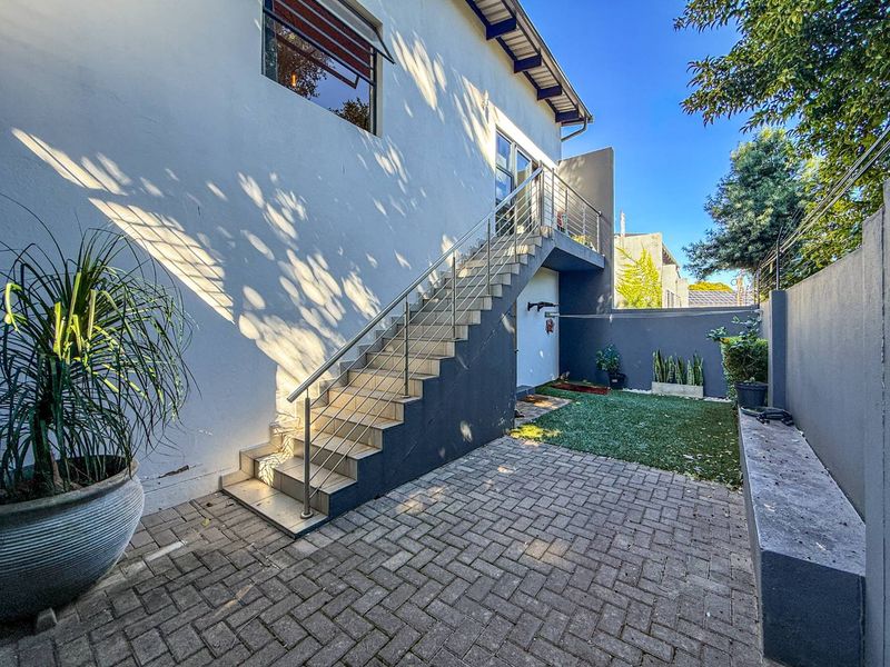 A well-maintained rear courtyard featuring a paved patio, artificial lawn, fixed concrete bench, and an external staircase with a modern stainless steel balustrade.
