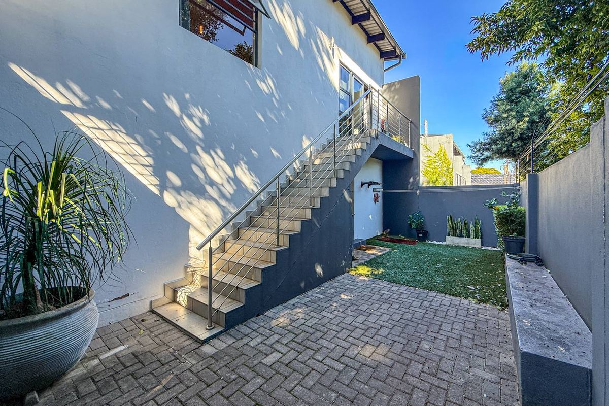 A well-maintained rear courtyard featuring a paved patio, artificial lawn, fixed concrete bench, and an external staircase with a modern stainless steel balustrade.