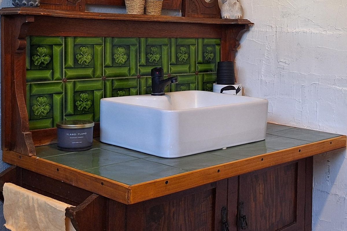 A vintage-style bathroom vanity featuring a white ceramic vessel sink, green decorative tile backsplash, and a wooden cabinet base.