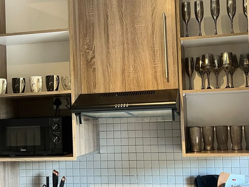 A modern kitchen area featuring wood-look cabinetry, a black granite countertop, a square mosaic tile backsplash, and a built-in electric hob with extractor fan.