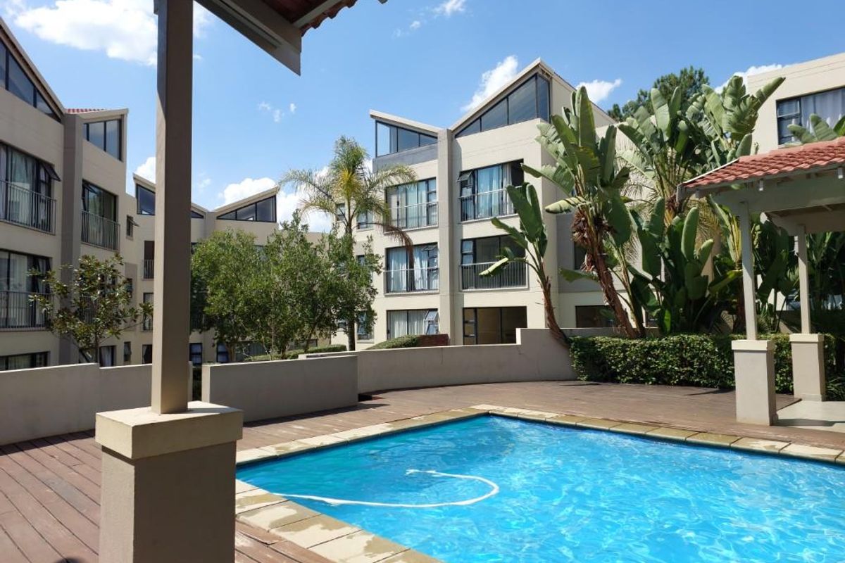 A communal swimming pool area featuring a wooden deck, covered patio structure, and landscaped garden set within a modern apartment complex.