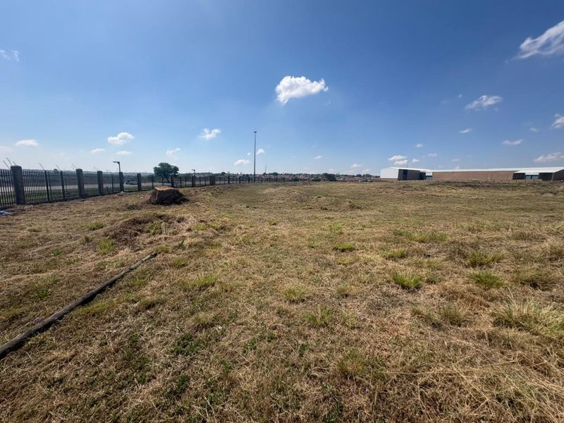 A wide-angle view of a vacant commercial land plot featuring perimeter fencing and security spikes under a clear blue sky.