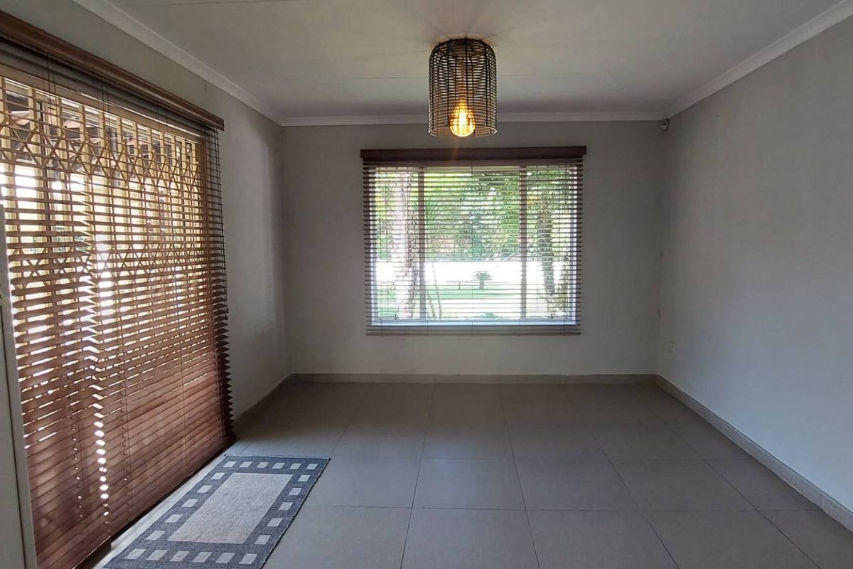 A bright, empty living room featuring neutral tiled flooring, a sliding glass door with security gate, and natural light through venetian blinds.
