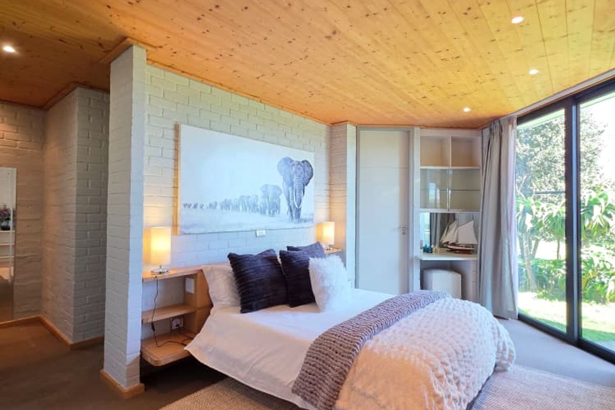 A well-lit bedroom featuring a warm timber-clad ceiling, painted brick walls, built-in bedside shelving, and large sliding glass doors providing garden access.