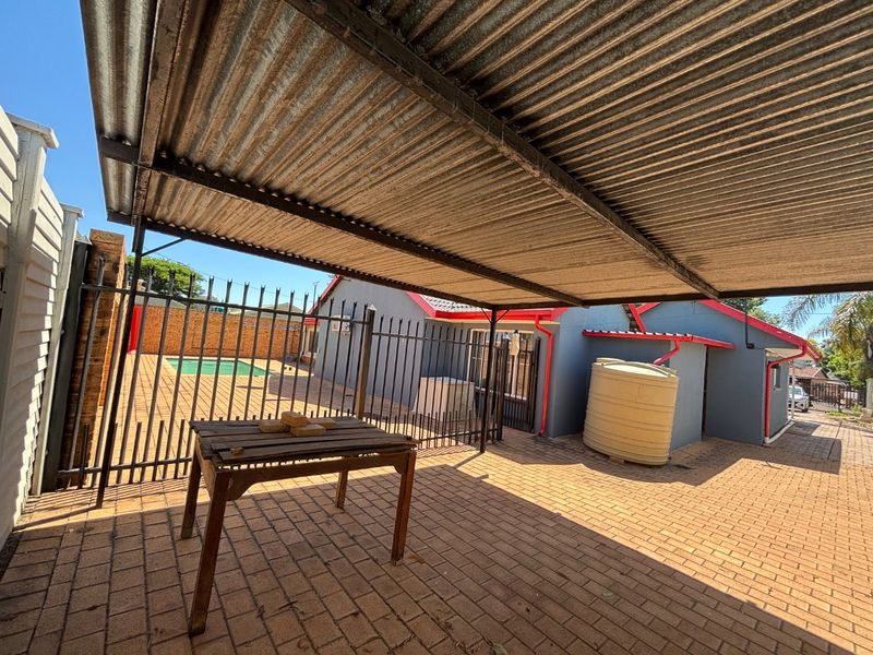 Covered outdoor patio area featuring paved flooring, a corrugated iron roof structure, and direct access to the swimming pool and garden area.