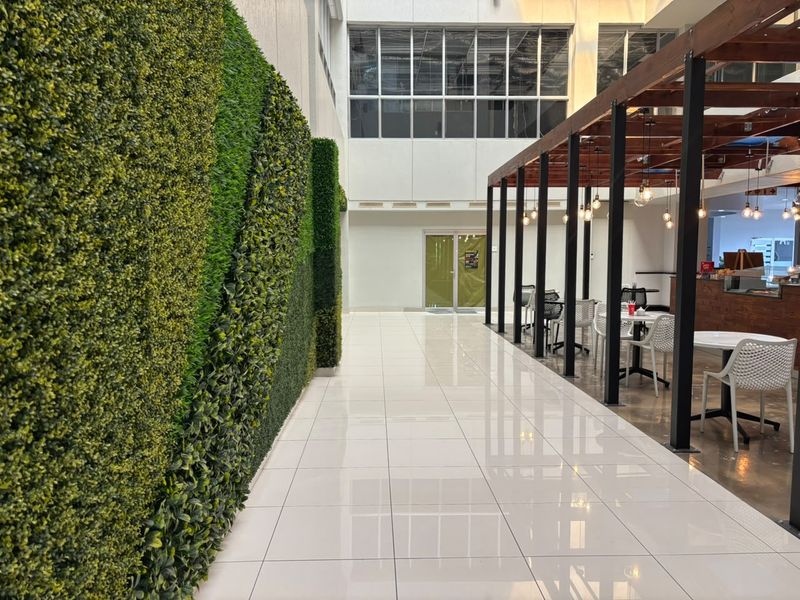 Modern open-plan office breakout area featuring a striking artificial green wall, polished tile flooring, and a timber pergola structure with pendant lighting.