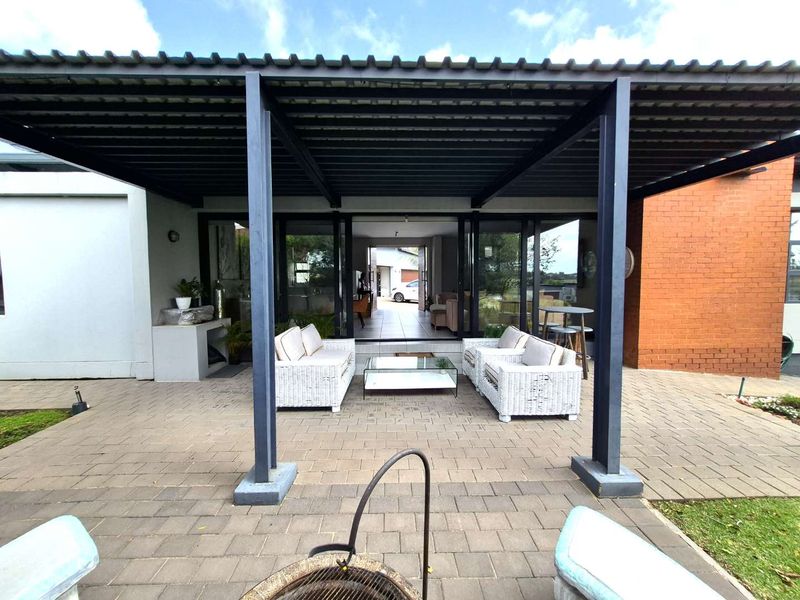 A spacious covered patio featuring a metal pergola, paved flooring, and a built-in braai, providing seamless access to the interior through sliding glass doors.