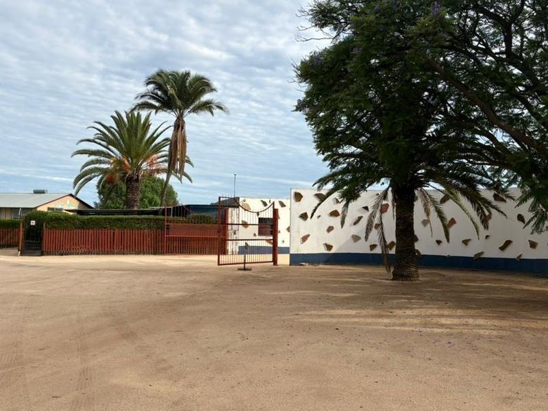 Front view of the commercial property featuring a large open gravel area, perimeter wall, and gated entrance with mature trees.