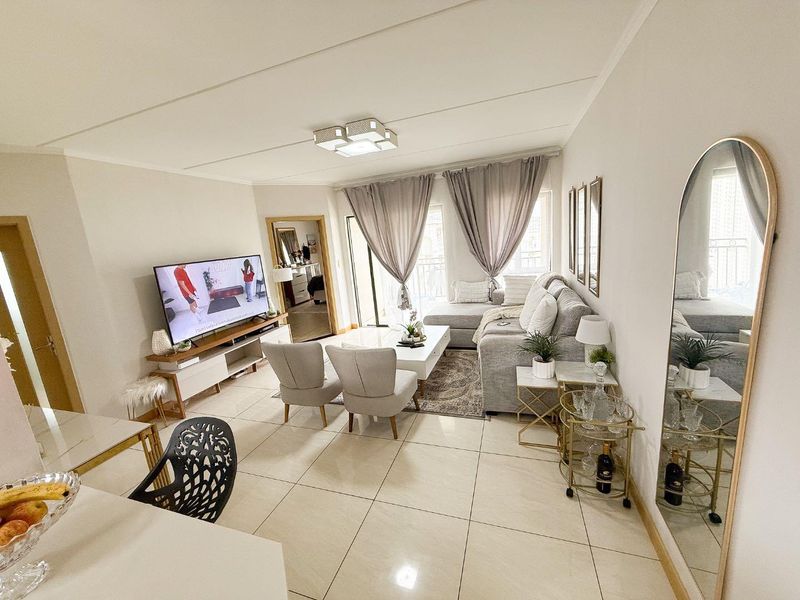 A bright and well-maintained living room featuring large format floor tiles, neutral walls, and direct access to a balcony through sliding glass doors.