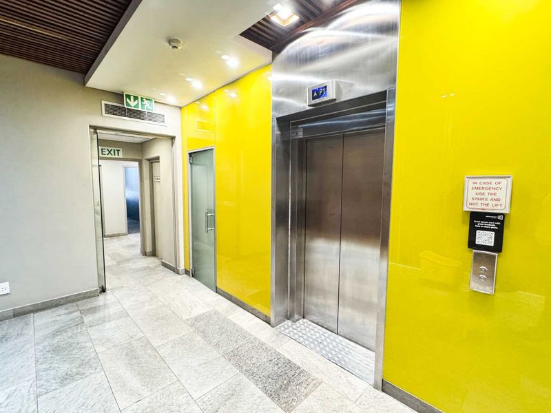 A modern commercial hallway featuring a stainless steel elevator, bright yellow wall paneling, and clean tiled flooring with clear emergency exit access.
